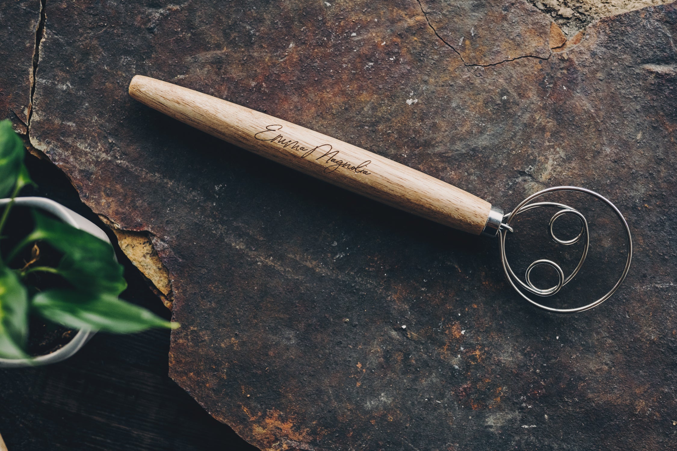 Wooden whisk handle with metal whisk head and Emma Magnolia embossed on a dark stone surface
